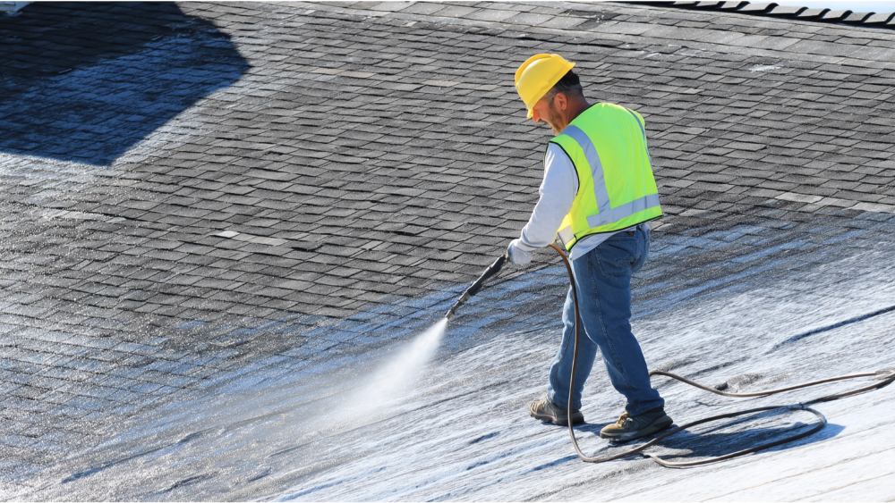 Worker wearing a safety vest and helmet pressure washing a shingle roof to remove buildup, spraying in wide strokes.