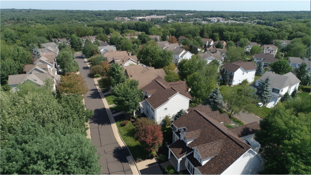 Aerial view of a residential neighborhood with various roof colors and styles, surrounded by trees and green landscapes.