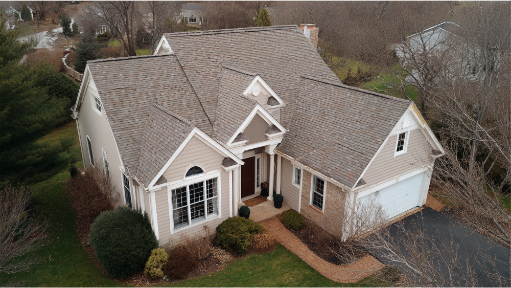 Two-story beige suburban home with a gray asphalt shingle roof, white trim, and a brick walkway leading to the entrance.