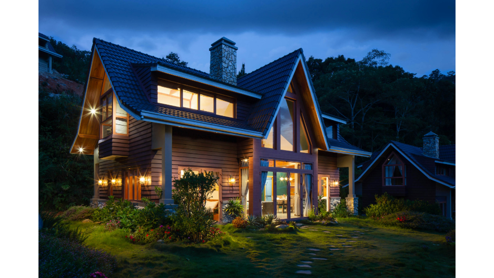 Modern mountain-style home with a steep pitched roof, stone chimney, and large front windows illuminated at dusk.