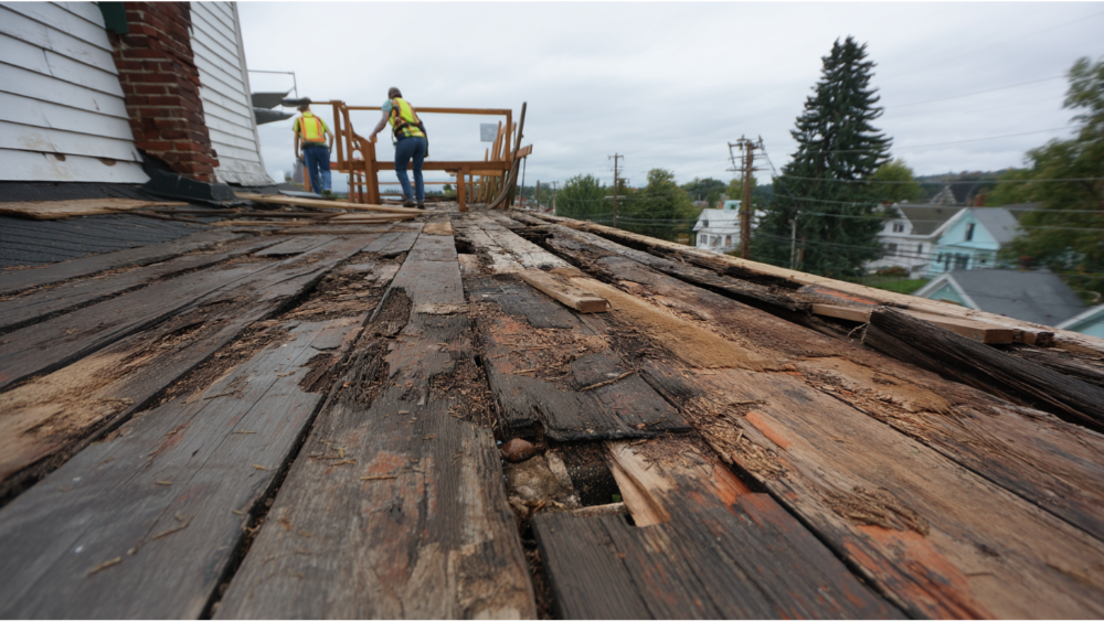 Old, weathered roof decking with extensive water damage and rot, with workers in the background preparing for repair."