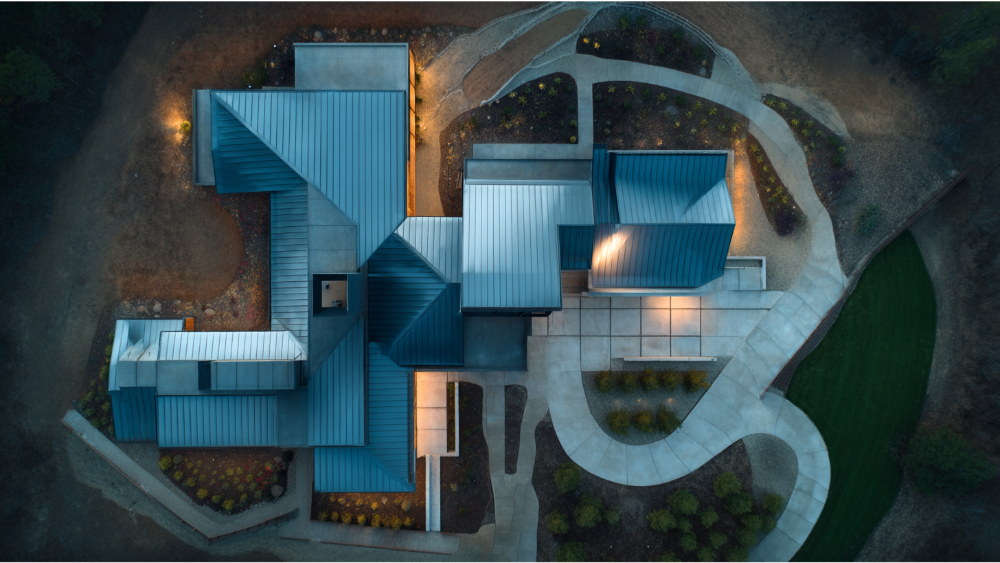 Drone shot of a modern home with a complex metal roofing layout, lit by exterior lighting in the early evening